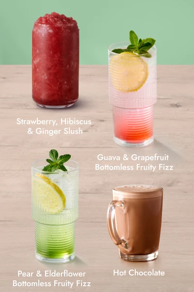 Five different drinks on a wooden surface with a green background: Hot Chocolate, Guava & Grapefruit Bottomless Fruity Fizz, Strawberry & Hibiscus Ginger Slush, Pear & Elderflower Bottomless Fruity Fizz, and a new Rolo Muggachino with whipped cream and chocolate drizzle.