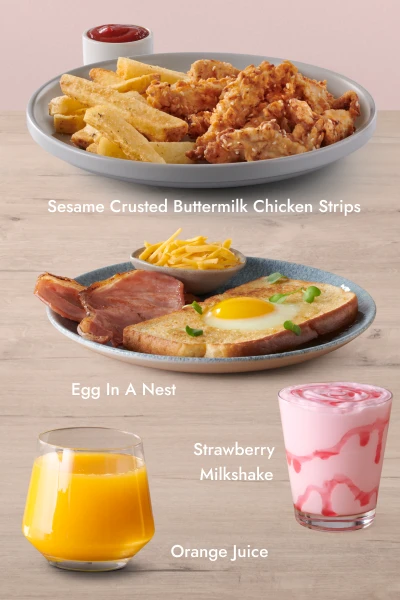 Three food and drink items on a wooden surface with a pink background: a plate of Sesame Crusted Buttermilk Chicken Strips with fries and ketchup, a glass of orange juice, and a plate of Egg In A Nest with grilled ham and a bowl of grated cheese.