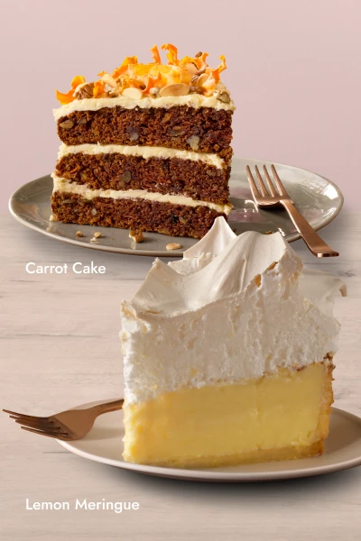 Two dessert slices on plates: a tall Lemon Meringue Slice with a fluffy meringue topping and creamy lemon filling, and a layered Carrot Cake Slice with cream cheese frosting and topped with nuts and carrot shavings.