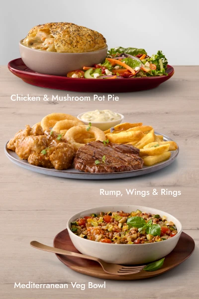Three plated meals on a wooden table: Chicken Mushroom Pot Pie with side salad, Rump steak with chicken wings, onion rings, fries, and dipping sauce, and a Mediterranean Veg Bowl with grains, vegetables, and herbs.