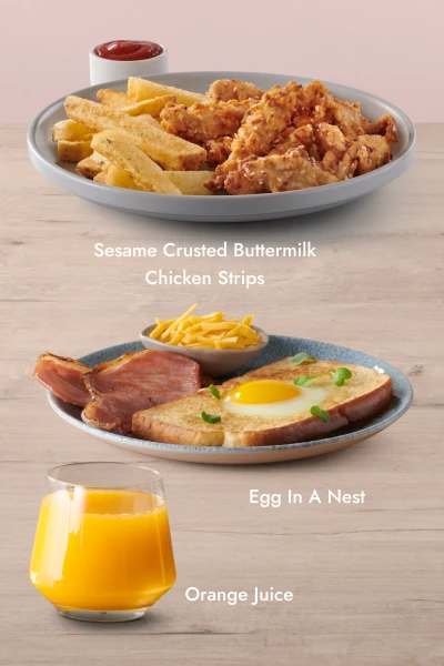 Three food and drink items on a wooden surface with a pink background: a plate of Sesame Crusted Buttermilk Chicken Strips with fries and ketchup, a glass of orange juice, and a plate of Egg In A Nest with grilled ham and a bowl of grated cheese.