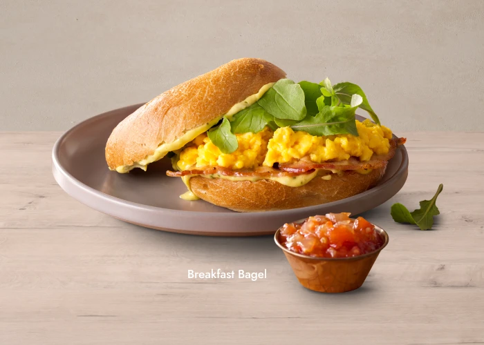 A breakfast bagel sandwich filled with scrambled eggs, crispy bacon, and fresh arugula, served on a ceramic plate with a small side cup of tomato relish.