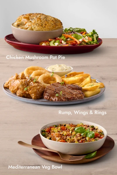 Three plated meals on a wooden table: Chicken Mushroom Pot Pie with side salad, Rump steak with chicken wings, onion rings, fries, and dipping sauce, and a Mediterranean Veg Bowl with grains, vegetables, and herbs.